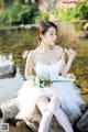 A woman in a white dress sitting on a rock by the water.