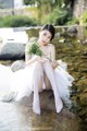 A woman in a white dress sitting on a rock by a river.