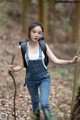 A woman in overalls and a backpack holding a stick in the woods.